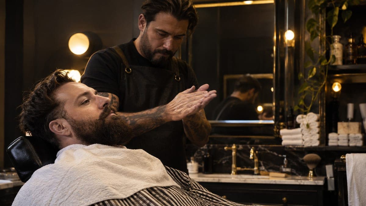 A barber warming beard balm between his palms before combing it through a customer's beard