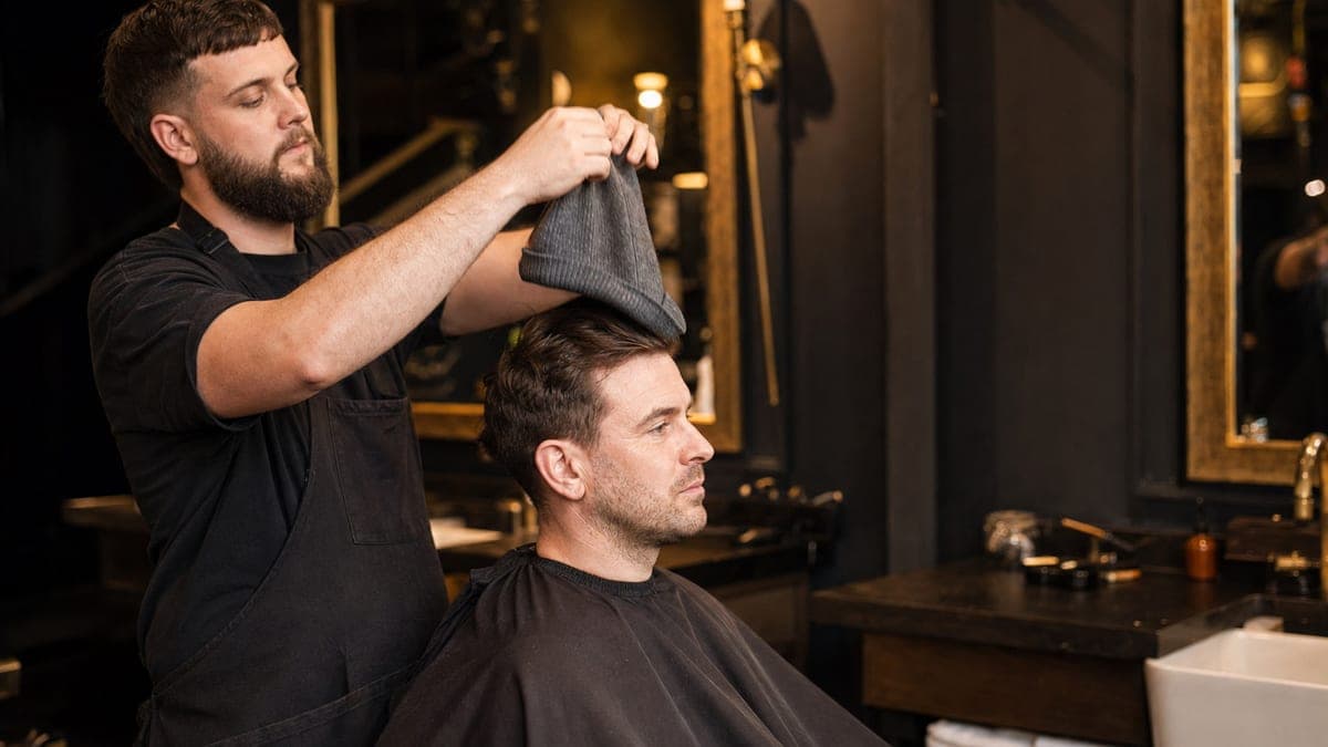 A barber lifting a grey knitted beanie straight up off the head of a man seated in the chair, wearing a black cape